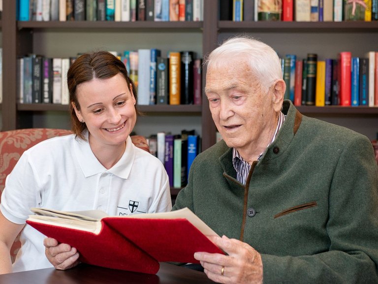 im Wohnzimmer: ein Bewohner liest aus einem Buch - eine junge Pflegerin sieht ond hört zu